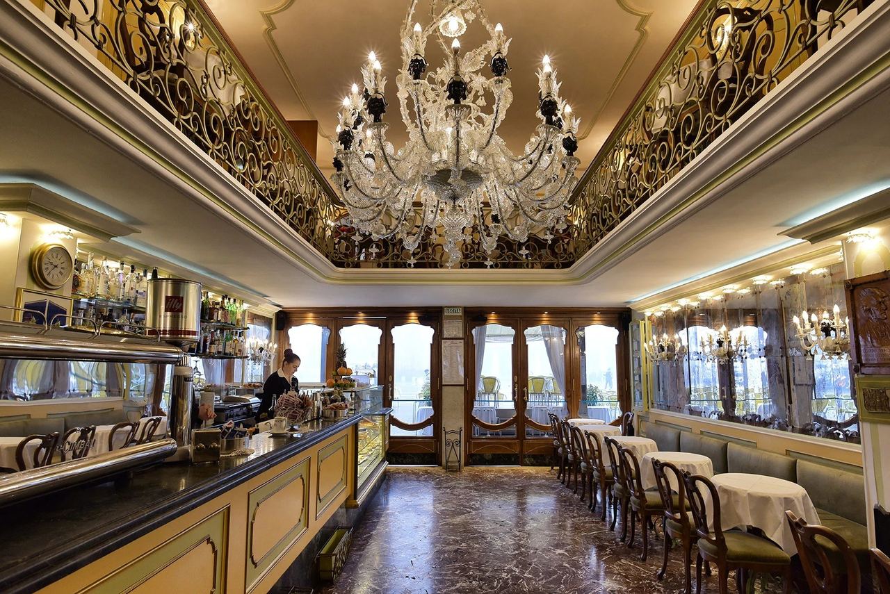 The mirrored room of Caffè Lavena under its Murano chandelier and wrought-iron gallery
